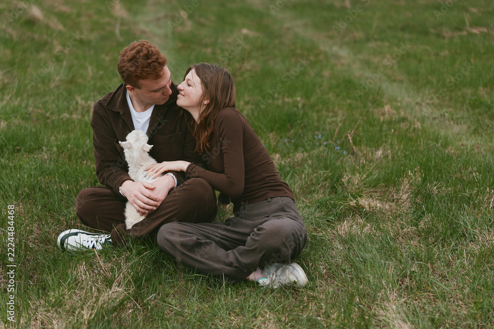 Fototapeta premium Couple sitting on grass in a park, sharing a tender kiss while holding a small white dog, a candid moment of affection and companionship in a natural setting with warm daylight