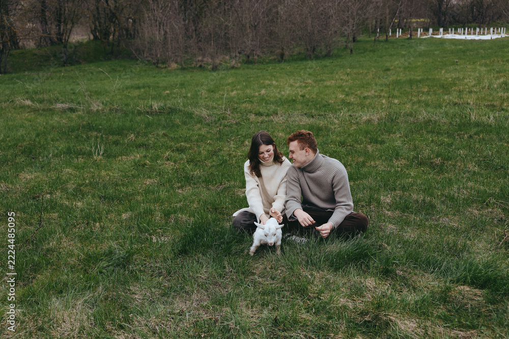 Fototapeta premium Couple with a white goat kid in a grassy field, sharing a gentle moment. Sunlit rural scene conveys lifestyle and affection, natural joy, and relaxed, candid connection between people and animals.