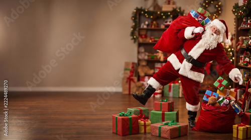 Funny Santa Claus in red costume carrying sack full of gifts tripping over Christmas presents on wooden floor in decorated living room with holiday decoration and gift boxes