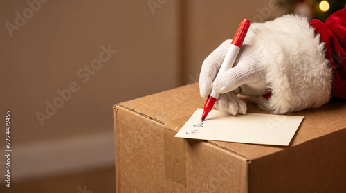 Santa Claus hand wearing white glove writing on gift tag with red marker on cardboard box, Christmas gift delivery and holiday preparation concept