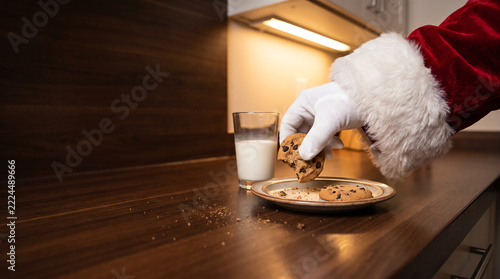 Santa Claus hand in white glove taking chocolate chip cookie from plate with glass of milk on wooden kitchen counter, Christmas eve tradition concept