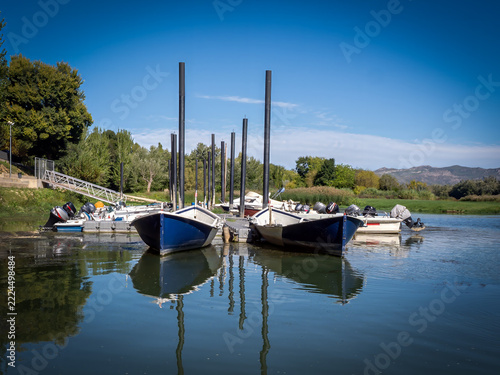 A peaceful row of boats is docked along a quiet pier, perfectly reflecting on the calm and still water of a scenic lake under a clear blue sky