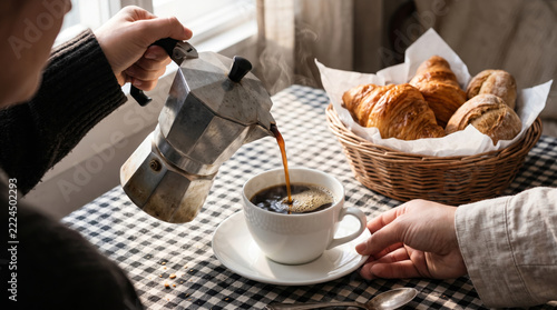 Pouring hot coffee from a moka pot into a cup with fresh croissants
