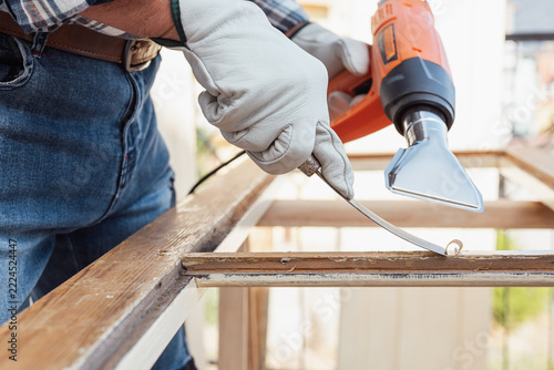 Adult carpenter wearing protective leather gloves, using a heat gun and a wood scraper to remove paint from an old wooden window. Construction industry, carpentry. Restoration. 