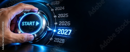 Human hand turning glowing metallic dashboard dial pointing precisely towards the glowing futuristic year twenty twenty seven typography against dark abstract background