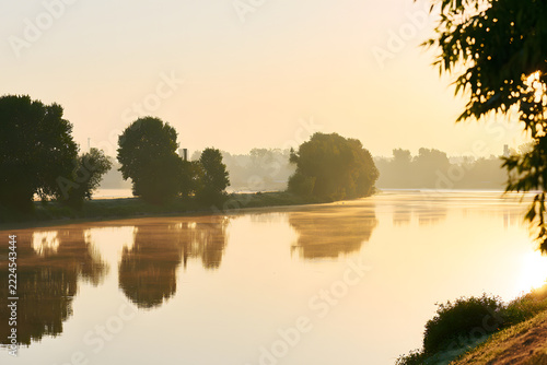 Golden sunrise light illuminates a serene river with misty surface, reflecting silhouetted trees on the banks. A tranquil and peaceful nature landscape at dawn.