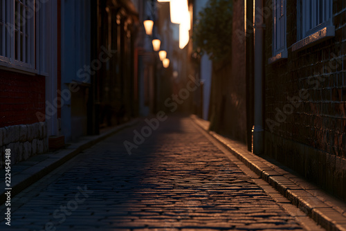 Narrow, atmospheric cobbled alley at night, lit by warm streetlights casting long shadows on the old stone pavement, creating a mysterious mood.