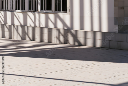 Sunny architectural scene with strong geometric shadows on a white building wall, stone base, and tiled ground, creating abstract patterns and depth.