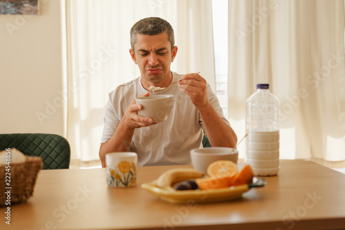 Man with a disgusted grimace holding a bowl and spoon while eating tasteless breakfast at a kitchen table.
