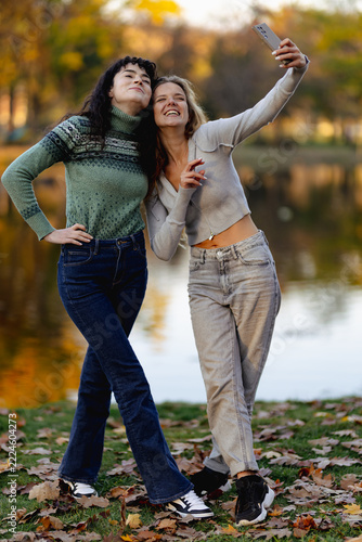 Two friends take a cheerful selfie together by a picturesque lake during autumn, surrounded by falling leaves and vibrant colors of the season