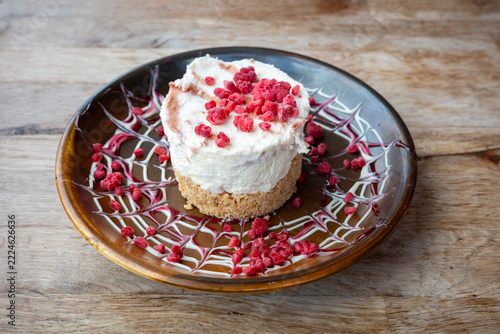 White chocolate and strawberry cheesecake on a brown dish decorated with white and red sauce and fresh fruit, on a rustic wooden table.