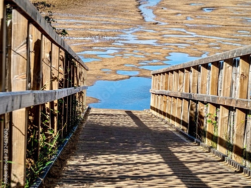 Perspective view looking down a wooden coastal walkway towards tidal mudflats and shallow blue water
