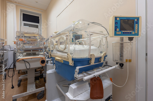 An empty neonatal incubator with a transparent hood and control panel stands beside other incubators in a hospital room