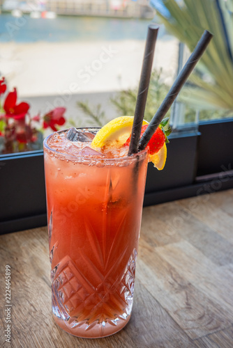 Strawberry lemonade iced drink with two paper straws and a slice of lemon in a tall glass on a wooden table by a window.