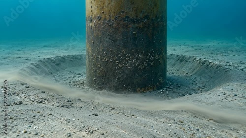 Underwater metal piling on sandy seabed, rippled sand and drifting sediment reveal marine corrosion beneath clear blue water