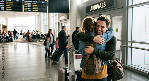 A smiling couple embraces warmly in an airport arrivals hall, reunited while other travelers walk past with luggage in the background.
