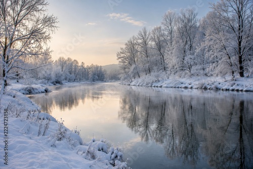 Quiet winter river reflecting bare frost covered trees along deep snowy banks