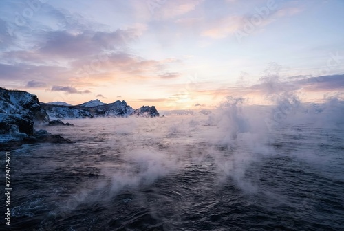 Sea smoke rising above cold ocean waters near snow-covered rocky shorelines under a morning sky.