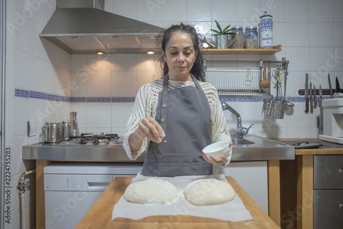 Mature woman sprinkling flour or salt over fresh raw dough balls in the kitchen