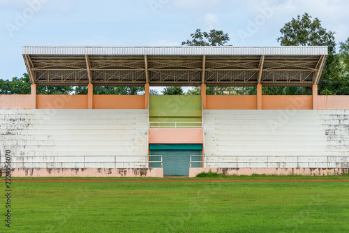 Empty Outdoor Sports Stadium Grandstand and Green Field at Remote Area. Front view of an rural covered stadium grandstand with white concrete seating, colorful walls, and a green grass sports field.