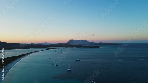Cinematic drone shot of the Sardegna coast at sunset featuring calm blue waters, white sand beaches, and distant mountains. Perfect for travel documentaries and wellness retreats