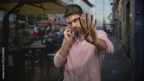 Fotografie Man with beard holding smartphone to ear, palm raised in stop gesture toward camera, standing at a street cafe sidewalk with tables and umbrellas; annoyance privacy