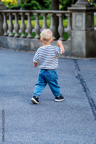 Little blonde toddler boy running away on pathway in green summer park back view, happy childhood concept
