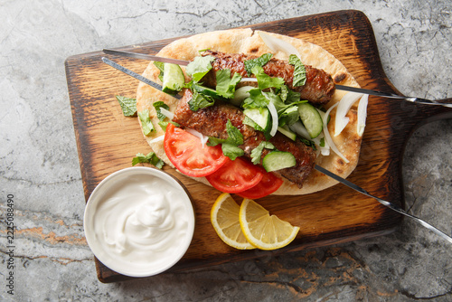 Grilled seekh kebab skewers with yogurt, naan bread and vegetables closeup on the wooden board. Horizontal top view from above