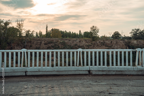 A serene view from a rural bridge captures the setting sun casting a warm glow over the landscape. The white railing frames a backdrop of trees and distant hills.