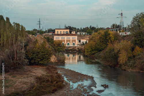 A prominent building stands by the river, surrounded by lush trees and set against a serene landscape. Power lines and infrastructure are visible in the background, adding contrast.
