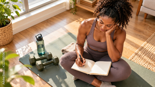 Young fit woman in activewear writing in a journal at home workout space with dumbbells and water bottle, perfect for fitness planning
