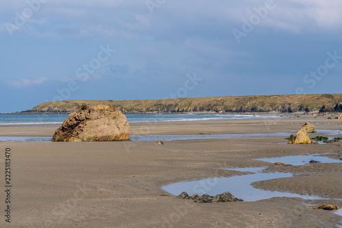 A large isolated rock on a wide sandy beach with shallow tidal pools and a calm sea near Porth Colmon on the Welsh coast. Natural coastal landscape with gentle waves, distant cliffs, and open space un