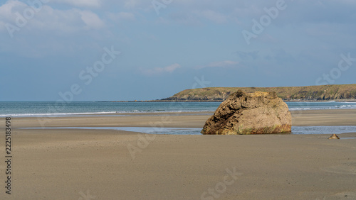 A large solitary boulder resting on a wide sandy beach with calm sea and distant coastal headland near Porth Colmon on the Welsh coast. Minimalist coastal scene with open space, gentle waves, and soft