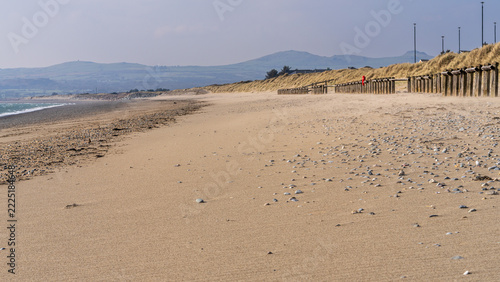 A wide sandy beach scattered with pebbles, backed by dunes and wooden groynes, stretching into the distance under clear daylight in Pwllheli on the Llŷn Peninsula. Calm coastal scene with strong leadi