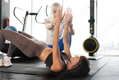 Two women performing partnered core exercise on gym mats with trainer guiding form during bright studio workout, with women, trainer, exercise, workout detail, showing fitness, core, training