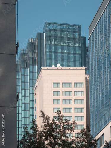 Layered city architecture featuring a beige building and modern blue glass skyscrapers behind autumn foliage.