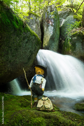 Vintage teddy bear dressed as traditional hiker with backpack and walking stick in front of forest waterfall