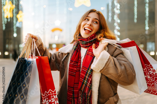Woman with red scarf stands in front of shopping center, holding her shopping bags. Smiling after her shopping trip, she enjoys Christmas atmosphere. Concept of holidays, gift-giving, and enjoyment.