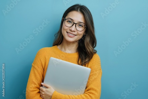 Portrait of young woman student smiles warmly while holding laptop on blue background. She wears yellow sweater and round glasses