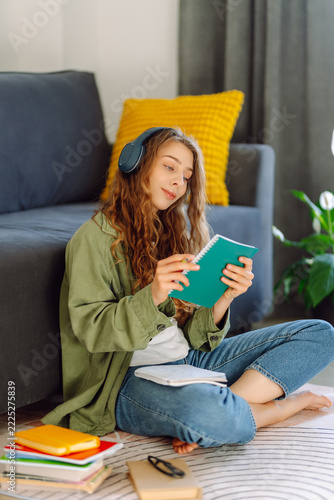 Woman sits at home near sofa, wearing wireless headphones and holding book. Young student takes notes while sitting in sunlit living room. Concept of education, leisure, and study.