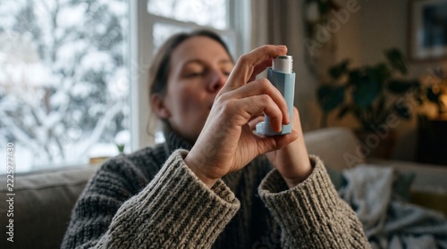 Close-up of a person wearing a cozy knit sweater using a blue inhaler for asthma treatment indoors near a snowy window during winter.