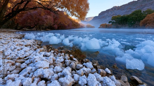 Low-angle view of ice floes along a rocky lakeshore, surrounded by changing autumn foliage and dramatic mountains in a cold highland landscape of Pakistan.