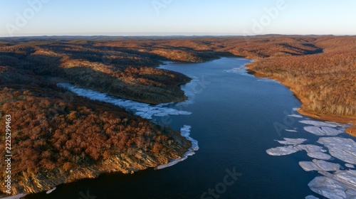 Panoramic aerial view of a tranquil mountain lake in Pakistan, surrounded by changing autumn foliage, rugged wilderness, and floating ice.