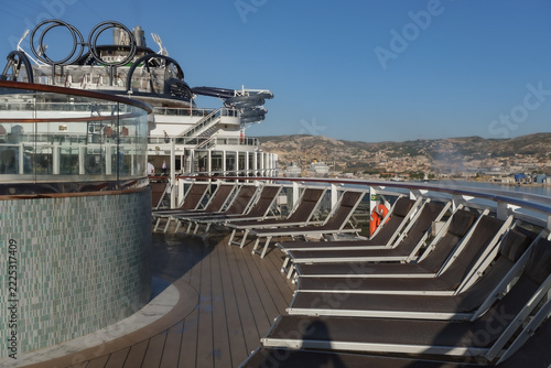 Pooldeck or swimmingpool with sun deck on modern cruiseship cruise ship liner in port with sun loungers ready for cruising to Mediterranean on sunny day with city skyline in background