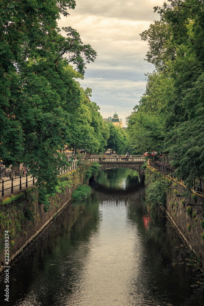 Fototapeta premium Uppsala, Sweden - 20.06.2026: Tranquil urban canal lined with lush green trees and a stone bridge under an overcast sky in Luthagen