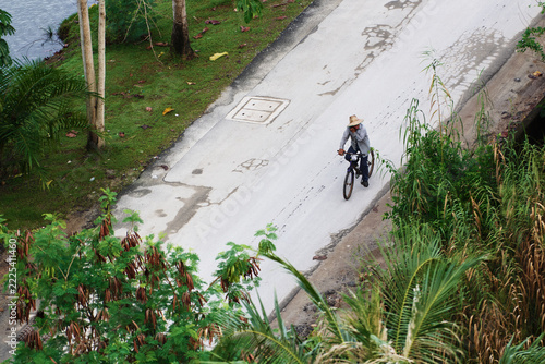 A lone cyclist pedals a bicycle along a quiet concrete road flanked by tropical plants and grass, conveying calm, everyday life, travel and outdoor activity in a rural or suburban setting.