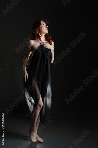Red haired woman in black sheer fabric, standing barefoot against dark studio background with dramatic side lighting