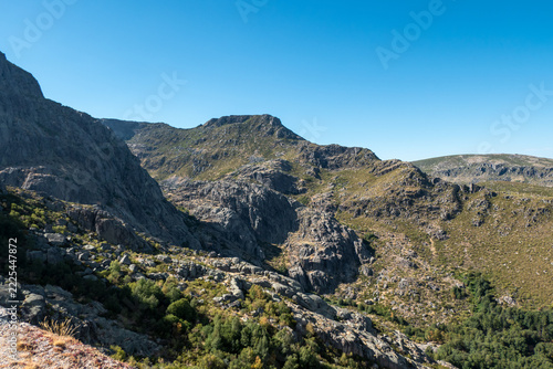 Paisagem montanhosa e rochosa da Serra da Estrela sob um céu azul limpo, com encostas de granito, vegetação rasteira e pequenos bosques nos vales em Portugal