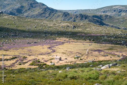 Pequena capela de pedra isolada na vasta paisagem montanhosa da Serra da Estrela, envolta por urze roxa, rocha granítica e vegetação rasteira em Portugal