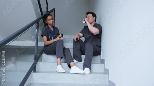 Two healthcare workers in gray scrubs sitting on stairs with takeaway coffee cups. Male and female colleagues talking during a break. Tired after hard day.